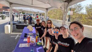 IMG 4775 Group of seven people in black T-shirts with BeakerBox Clemson Science Outreach on front, standing behind a purple table at a street fair.