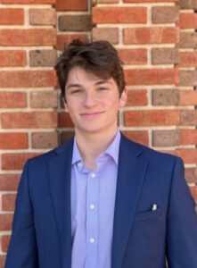 Male in Navy blazer with blue shirt standing in front of brick background.