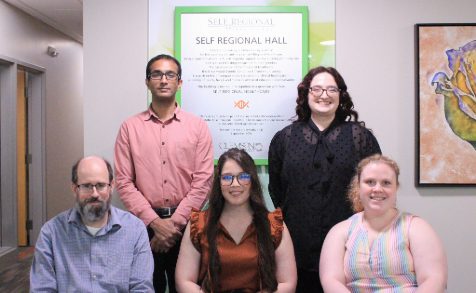 A photo of a man and a woman standing behind two seated women and one seated man in a hallway.
