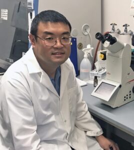 Zhicheng Dou sitting in the lab next to a microscope