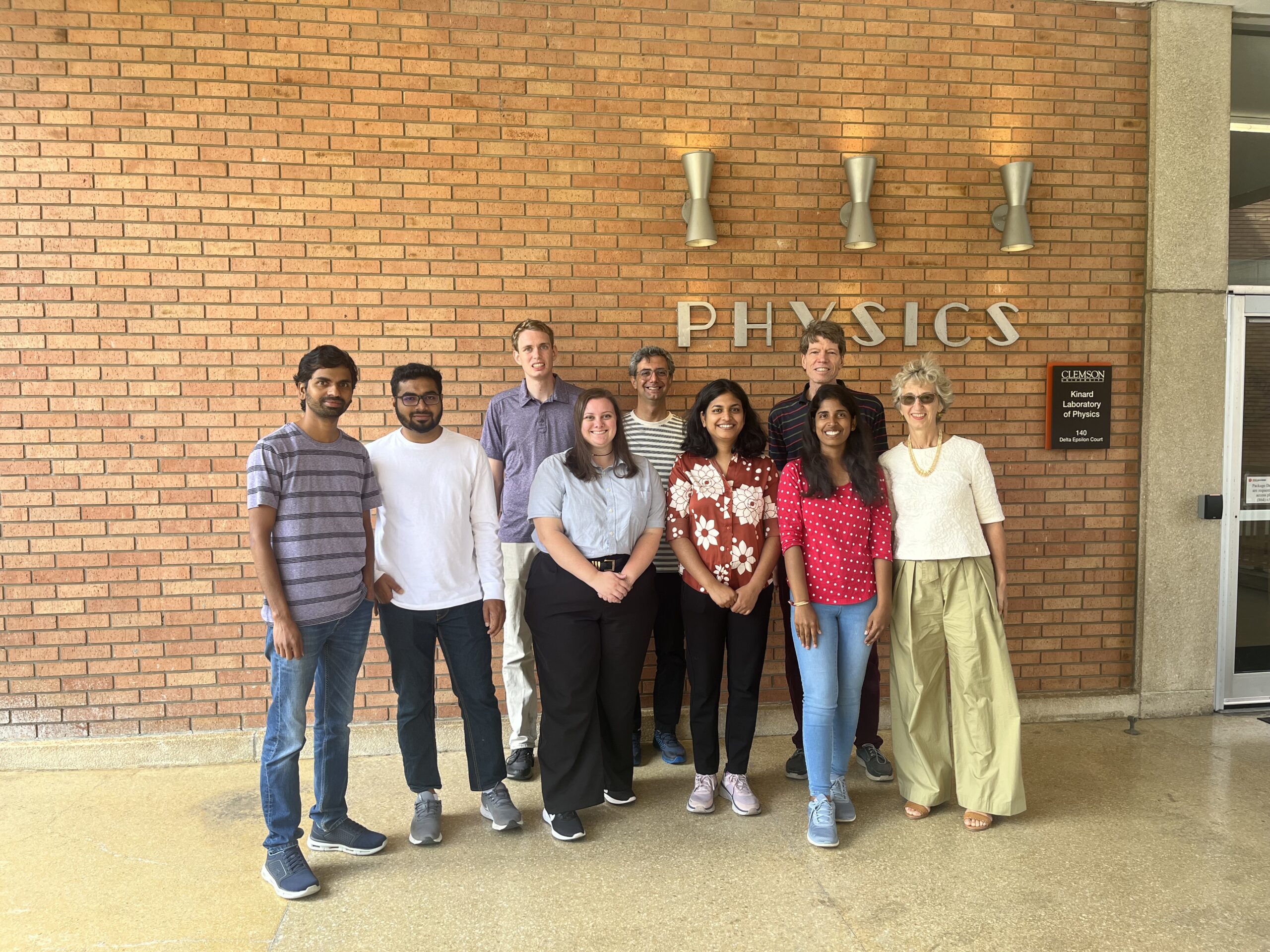 Group of nine people standing against an orange brick wall with PHYSICS in metal lettering behind them. A Clemson University sign on the right says Kinard Laboratory of Physics - 140 Delta Epsilon Court.