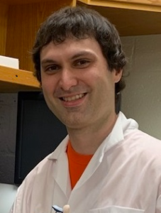 A man standing in a laboratory wearing an orange shirt underneath a white lab coat.