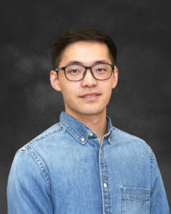 A man in front of a gray background wearing a blue denim button-up shirt and glasses.