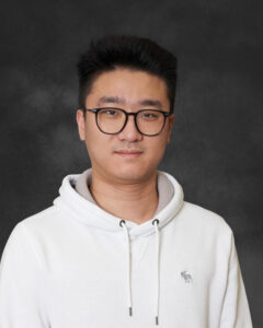 A man standing in front of a gray background wearing a white hoodie and glasses.
