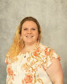 A woman in a white and pink floral dress with blonde hair against a grey background.