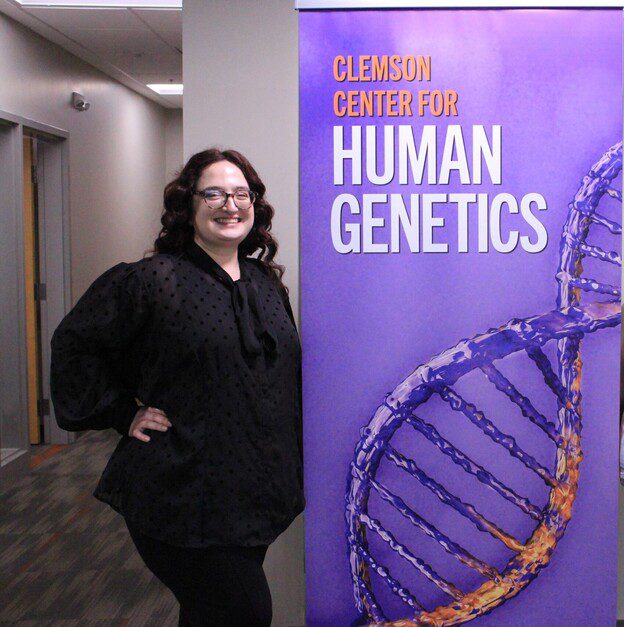 A woman with dark hair and in glasses dressed in black standing next to a purple banner that says Clemson Center for Human Genetics