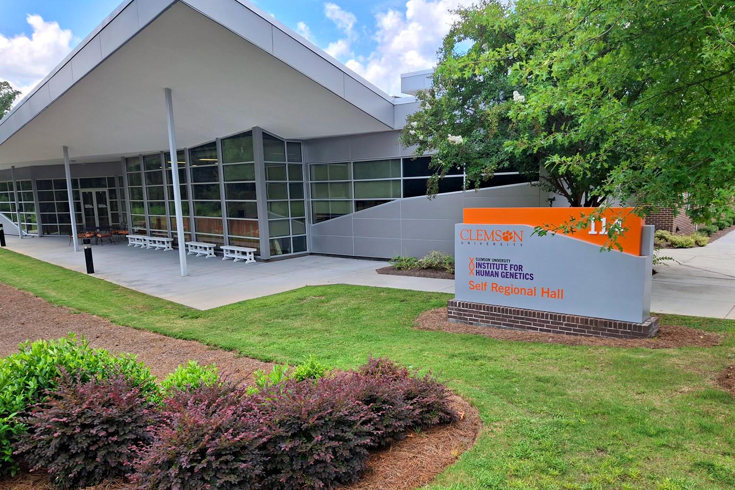 ihg homepage building Exterior of Clemson University’s Institute for Human Genetics at Self Regional Hall, featuring a modern gray building, orange Clemson signage, landscaped lawn, shrubs, and trees.