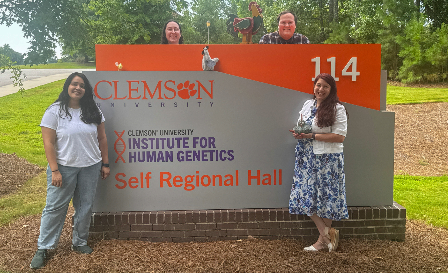 Four people pose around a Clemson University Institute for Human Genetics sign outside Self Regional Hall; two stand in front while two peek over the sign, with colorful bird decorations on top.